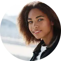 Personne souriante aux cheveux bouclés, vêtue d'une veste en jean, dans un décor extérieur ensoleillé avec un pont flou en arrière-plan, dégageant une atmosphère joyeuse