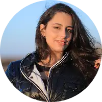 Femme souriante aux cheveux bruns vêtue d'une veste noire, debout à l'extérieur avec en arrière-plan une plage ensoleillée et un ciel bleu flous