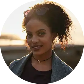 Une femme aux cheveux bouclés, portant des lunettes de soleil sur la tête, sourit doucement. Elle se trouve à l'extérieur, près de l'eau, au coucher du soleil, créant une ambiance chaleureuse et sereine