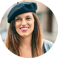 Une femme aux longs cheveux bruns sourit chaleureusement, coiffée d'un béret noir et vêtue d'un pull gris. Elle se tient debout dans une rue urbaine floue, dégageant une atmosphère joyeuse et détendue